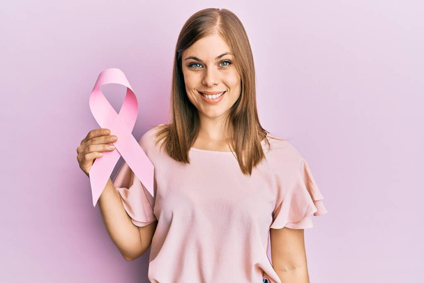 Beautiful caucasian woman holding pink cancer ribbon looking positive and happy standing and smiling with a confident smile showing teeth  - Foto, immagini