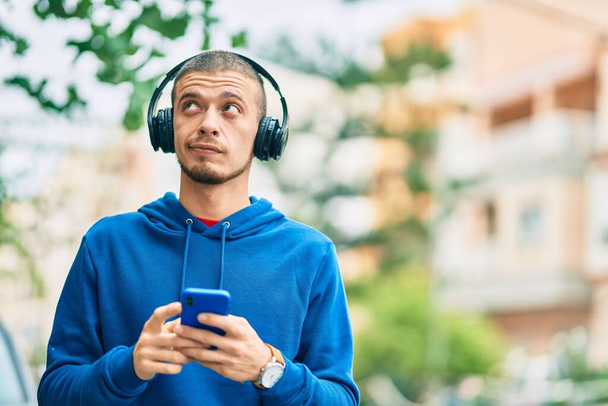 Mladý hispánec s vážným výrazem pomocí smartphonu a sluchátek ve městě. - Fotografie, Obrázek