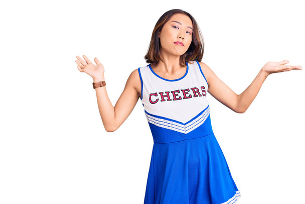 Młoda piękna Chinka w mundurze cheerleaderki, bez pojęcia i myląca ekspresja z podniesionymi rękami i rękami. koncepcja wątpliwości.  - Zdjęcie, obraz