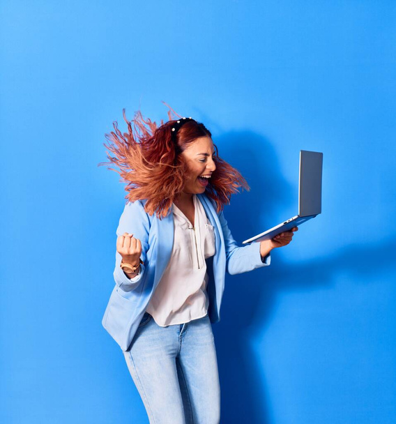 Young beautiful latin businesswoman smiling happy celebrating success. Jumping with smile on face using laptop over isolated blue background - Photo, Image