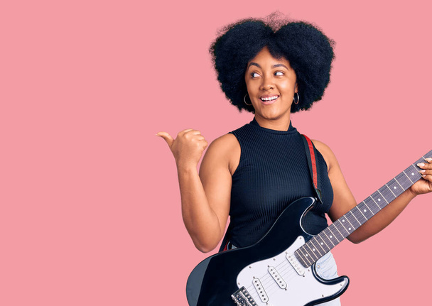 Jovem afro-americana tocando guitarra elétrica apontando polegar até o lado sorrindo feliz com a boca aberta  - Foto, Imagem
