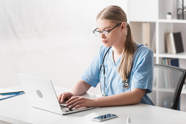 young veterinarian in glasses typing on laptop near smartphone on desk - Foto, Bild