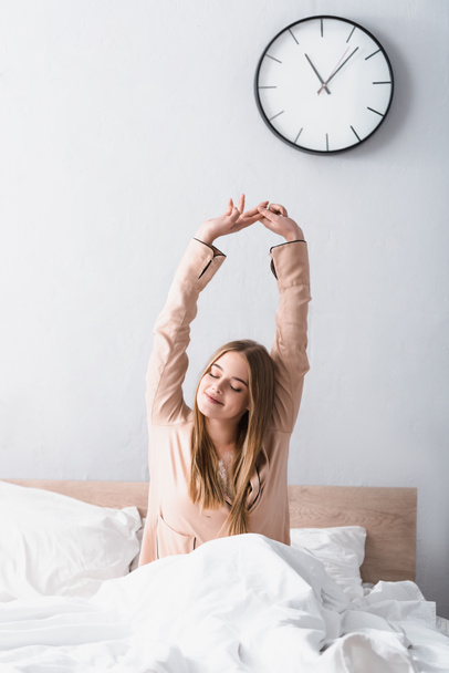 wakker en positief vrouw in satijnen pyjama stretching in bed - Foto, afbeelding