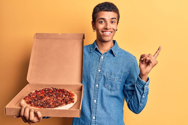 Νεαρός Αφροαμερικάνος που κρατάει το κουτί της πίτσας χαμογελώντας χαρούμενος δείχνοντας με το χέρι και το δάχτυλο στο πλάι  - Φωτογραφία, εικόνα