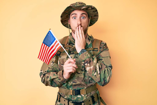 Giovane uomo caucasico che indossa l'uniforme dell'esercito mimetico che tiene la bandiera USA che copre la bocca con la mano, scioccato e spaventato per errore. espressione sorpresa  - Foto, immagini