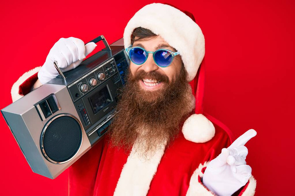 Handsome young red head man with long beard wearing santa claus costume and boombox smiling happy pointing with hand and finger to the side  - Fotó, kép
