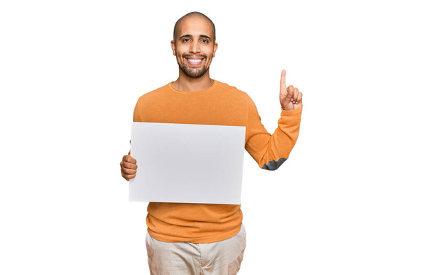 Взрослый латиноамериканец держит пустой баннер, удивленный идеей или вопросом, указывая пальцем со счастливым лицом, номер один  - Фото, изображение