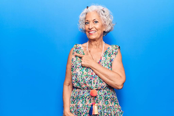 Ältere grauhaarige Frau in lässiger Kleidung lächelt fröhlich und zeigt mit der Hand und dem Finger zur Seite  - Foto, Bild