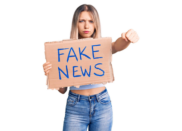 Молодая красивая блондинка с фальшивым баннером новостей раздражена и разочарована криками гнева, криками сумасшедшей от злости и поднятой рукой  - Фото, изображение