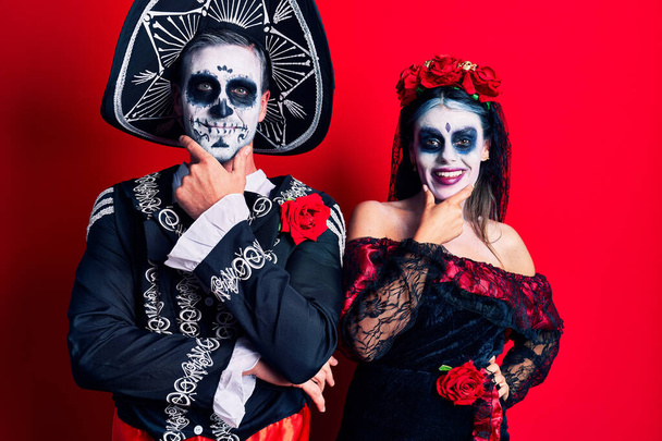 Young couple wearing mexican day of the dead costume over red looking confident at the camera smiling with crossed arms and hand raised on chin. thinking positive.  - Foto, imagen
