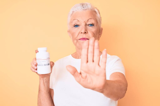 Старша красива жінка з блакитними очима і сірим волоссям тримає таблетки з відкритою рукою робить знак зупинки з серйозним і впевненим виразом, жест захисту
  - Фото, зображення