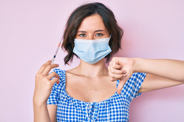 Young beautiful woman wearing medical mask holding syringe with angry face, negative sign showing dislike with thumbs down, rejection concept  - Photo, Image