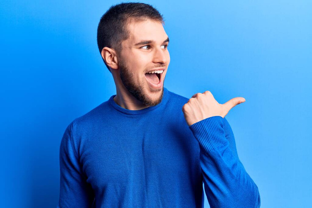 Young handsome man wearing casual sweater pointing thumb up to the side smiling happy with open mouth  - Fotografie, Obrázek