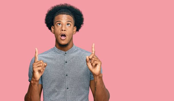 Hombre afroamericano con pelo afro vistiendo ropa casual asombrado y sorprendido mirando hacia arriba y señalando con los dedos y los brazos levantados.  - Foto, Imagen