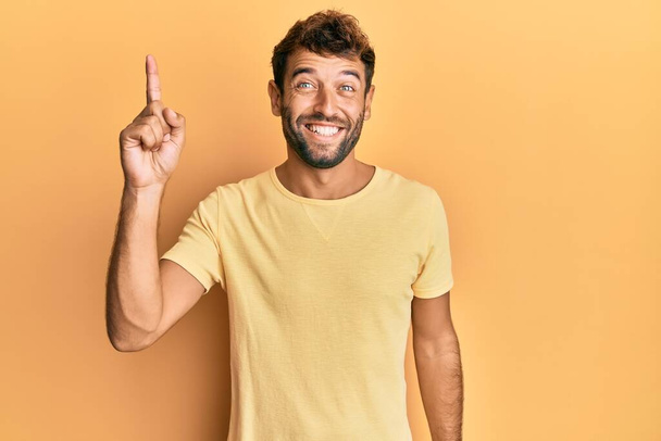 Handsome man with beard wearing casual yellow tshirt over yellow background pointing finger up with successful idea. exited and happy. number one.  - Photo, Image