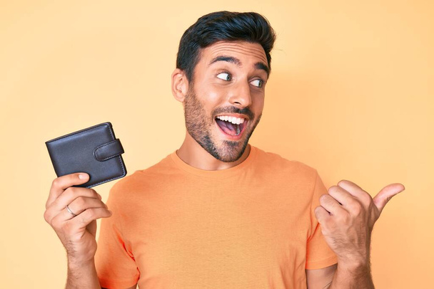 Молодий іспаномовний чоловік тримає шкіряний гаманець, вказуючи великий палець в сторону усміхнений щасливий з відкритим ротом
  - Фото, зображення