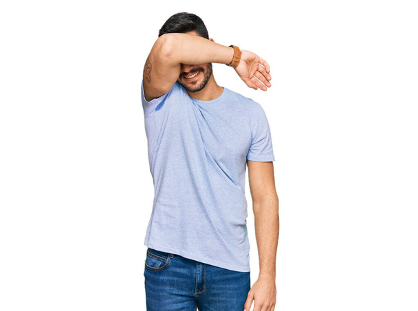 Young hispanic man wearing casual clothes covering eyes with arm smiling cheerful and funny. blind concept.  - 写真・画像