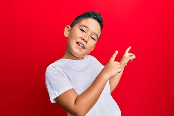 Günlük beyaz tişört giyen, gülümseyen ve kameraya bakan, iki eli ve parmağı yanda olan küçük İspanyol çocuk..  - Fotoğraf, Görsel