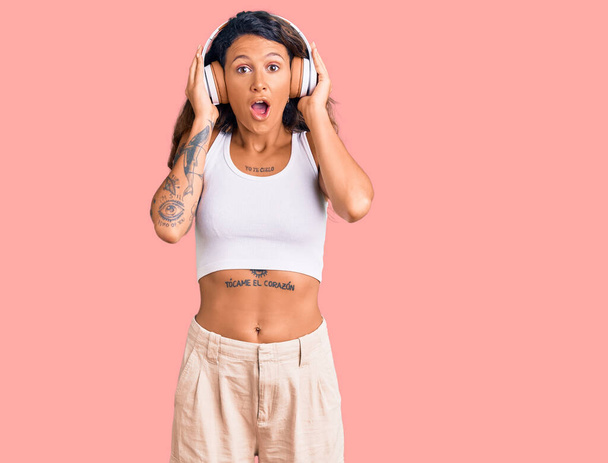 Młoda Latynoska kobieta z tatuażem słuchająca muzyki przy użyciu słuchawek przerażona i zdumiona otwartymi ustami dla zaskoczenia, niedowierzanie twarz  - Zdjęcie, obraz