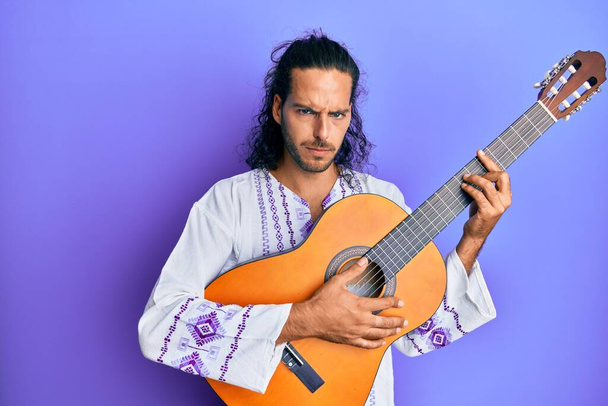 Mladý pohledný muž s dlouhými vlasy hrající na klasickou kytaru skeptický a nervózní, zamračený kvůli problému. negativní osoba.  - Fotografie, Obrázek