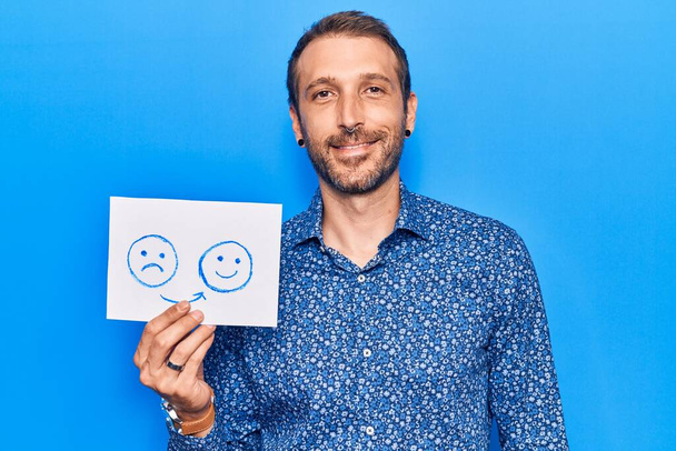 Fiatal jóképű férfi tartja szomorú boldog érzelem papír néz pozitív és boldog álló és mosolygó magabiztos mosollyal mutatja fogak  - Fotó, kép