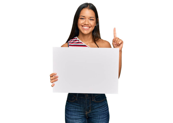 Красивая латиноамериканка держит пустой баннер, удивленный идеей или вопросом, указывая пальцем со счастливым лицом, номер один  - Фото, изображение