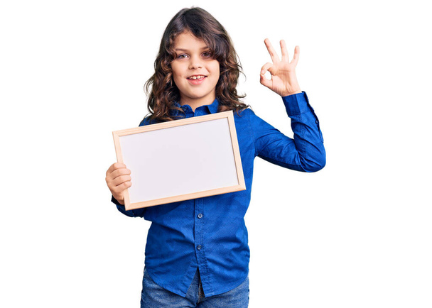 Cute Latynoski dziecko z długimi włosami trzymając puste białe tablice robi ok znak palcami, uśmiechnięty przyjazny gest doskonały symbol  - Zdjęcie, obraz