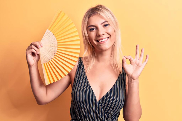 Junge schöne blonde Frau mit Handfächer, der über isoliertem gelben Hintergrund steht und ok Zeichen mit den Fingern macht, freundlich lächelnd, hervorragendes Symbol gestikulierend - Foto, Bild