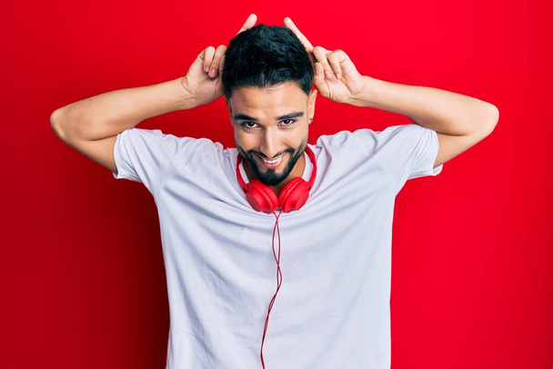 Young man with beard listening to music using headphones posing funny and crazy with fingers on head as bunny ears, smiling cheerful  - Valokuva, kuva