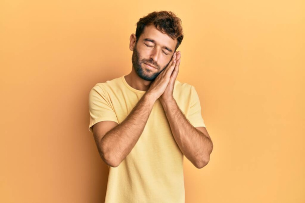Handsome man with beard wearing casual yellow tshirt over yellow background sleeping tired dreaming and posing with hands together while smiling with closed eyes.  - Fotó, kép