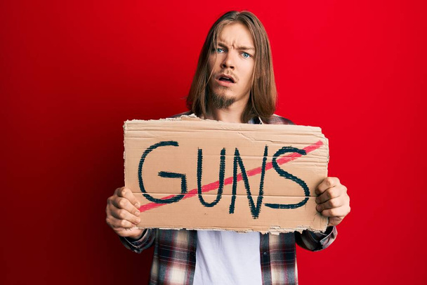 Handsome caucasian man with long hair holding no guns warning banner in shock face, looking skeptical and sarcastic, surprised with open mouth  - Photo, Image