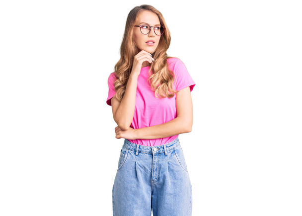 Young beautiful caucasian woman with blond hair wearing casual clothes and glasses with hand on chin thinking about question, pensive expression. smiling with thoughtful face. doubt concept.  - Фото, изображение