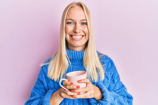 Chica rubia joven con suéter de invierno y bebiendo una taza de café caliente sonriendo con una sonrisa feliz y fresca en la cara. mostrando dientes.  - Foto, imagen
