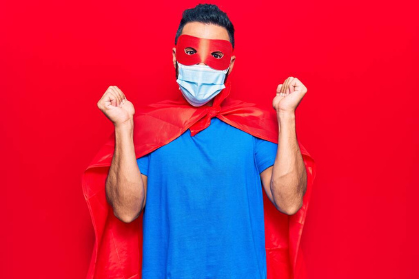 Молодий іспаномовний чоловік в костюмі супергероя і медичній масці кричить гордо, святкуючи перемогу і успіх дуже схвильований піднятою рукою
  - Фото, зображення