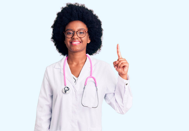 Genç Afro-Amerikalı kadın doktor önlüğü ve steteskop giyiyor. Bir numaralı parmağını gösterip işaret ederken kendine güvenen ve mutlu bir şekilde gülümsüyor..  - Fotoğraf, Görsel