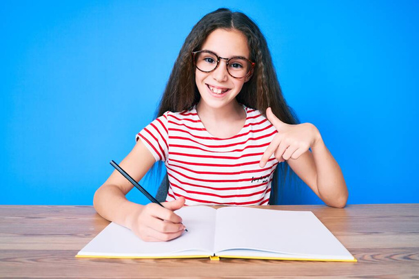 Nettes hispanisches Mädchen, das auf dem Tisch sitzt und Bücher schreibt, lächelt glücklich und zeigt mit Hand und Finger  - Foto, Bild