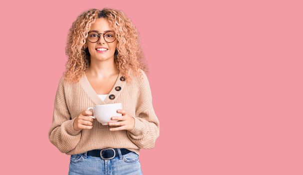 Mladá blondýnka s kudrnatými vlasy nosí brýle a pije šálek kávy vypadá pozitivně a šťastně stojící a usmívá se s sebevědomým úsměvem ukazující zuby  - Fotografie, Obrázek