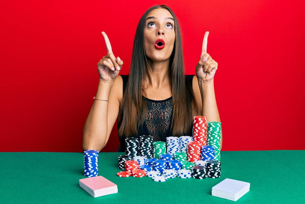 Genç İspanyol kadın masada oturmuş poker oynuyor. Şaşırmış ve şaşırmış bir şekilde yukarı bakıyor ve parmaklarıyla işaret ediyor ve kollarını kaldırıyor..  - Fotoğraf, Görsel