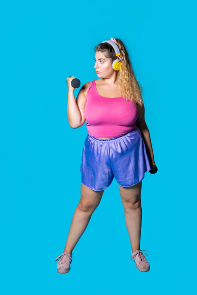 Plus size jonge zwarte vrouw gewicht tillen op blauwe achtergrond luisteren muziek dragen bluetooth draadloze hoofdtelefoon training - Foto, afbeelding
