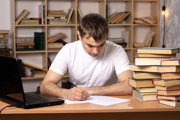 young male student writes an abstract at the table with books and laptop. European appearance. distance learning. preparation for exams. office worker is working. - Photo, Image