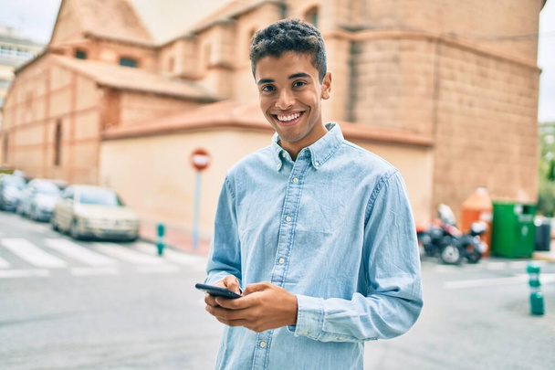Jovem latino sorrindo feliz usando smartphone andando na cidade. - Foto, Imagem