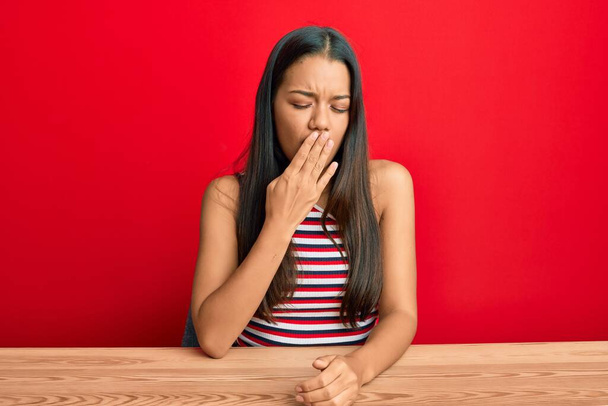 Beautiful hispanic woman wearing casual clothes sitting on the table bored yawning tired covering mouth with hand. restless and sleepiness.  - Foto, Bild