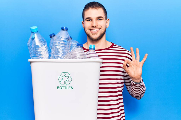 Młody przystojny mężczyzna recyklingu plastikowych butelek robi ok znak palcami, uśmiechając przyjazny gest doskonały symbol  - Zdjęcie, obraz