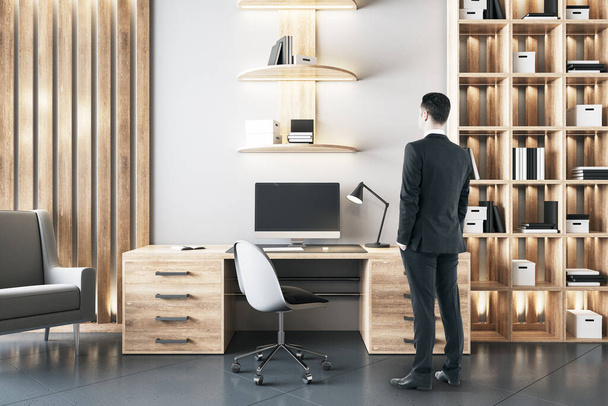 Homme d'affaires debout dans l'intérieur de bureau en bois ceo avec des ordinateurs sur la table et bibliothèque avec des livres. - Photo, image