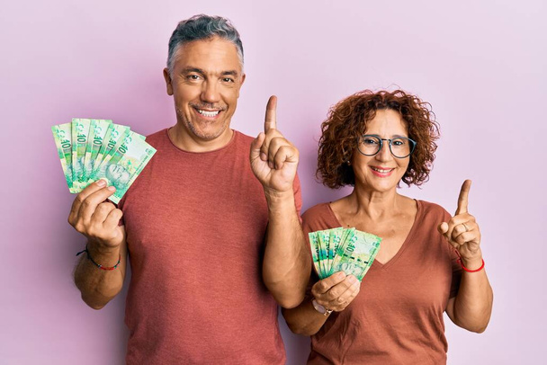 Красива пара середнього віку тримає південноафриканські банкноти 10 рандо, посміхаючись з ідеєю або питанням, спрямованим пальцем з щасливим обличчям, номер один
  - Фото, зображення