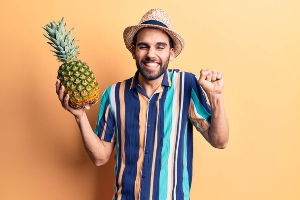 Young handsome man with beard wearing summer hat and shirt holding pineapple screaming proud, celebrating victory and success very excited with raised arm  - Foto, imagen