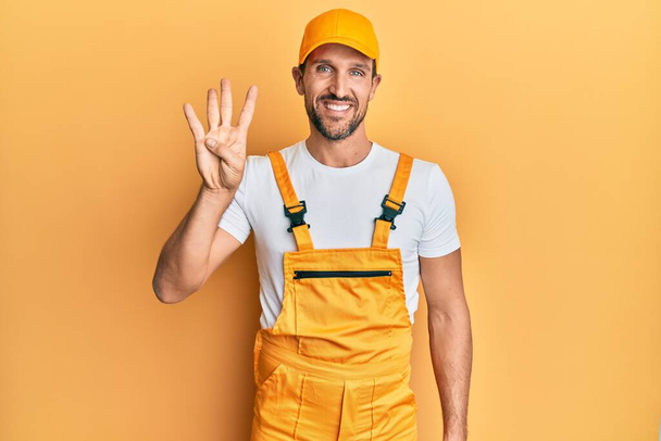 Jonge knappe man draagt klusjesman uniform over gele achtergrond tonen en wijzen met vingers nummer vier terwijl glimlachen zelfverzekerd en gelukkig.  - Foto, afbeelding