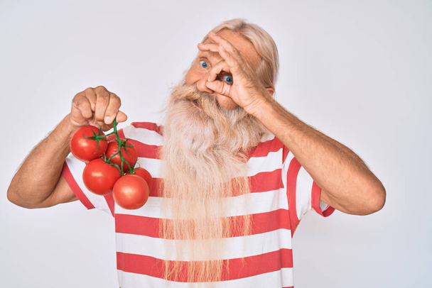 Starý starší muž s šedivými vlasy a dlouhými vousy drží freh zralá rajčata s úsměvem šťastný dělá ok znamení s rukou na oku dívá skrz prsty  - Fotografie, Obrázek