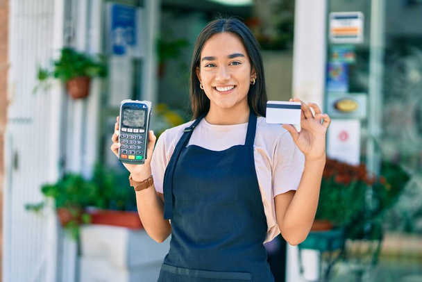 Joven tendero latino sonriendo feliz sosteniendo tarjeta de crédito y datáfono en la floristería - Foto, imagen