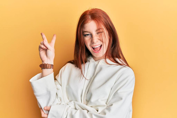 Young red head girl wearing casual sweatshirt smiling with happy face winking at the camera doing victory sign. number two.  - Valokuva, kuva